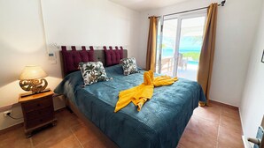 A bedroom with a bed, a nightstand, a lamp, and a view of the sea through a sliding glass door. - Villa Hereiti by ENJOY VILLAS / 3 bdrm AC /3 bath /private pool and front lagoon (PAPETOAI)