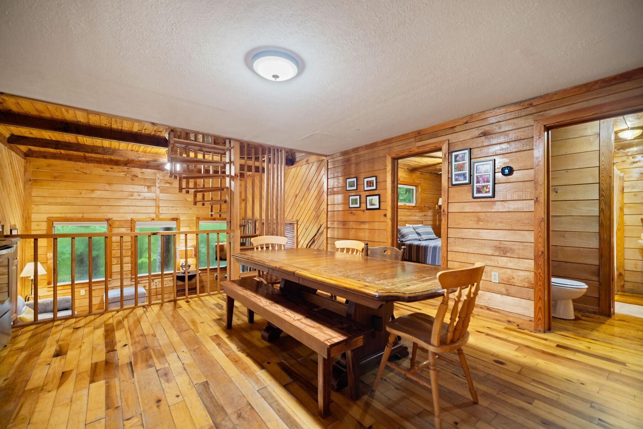 A wooden cabin interior with a large dining table, benches, a staircase, and a bathroom visible through an open door.