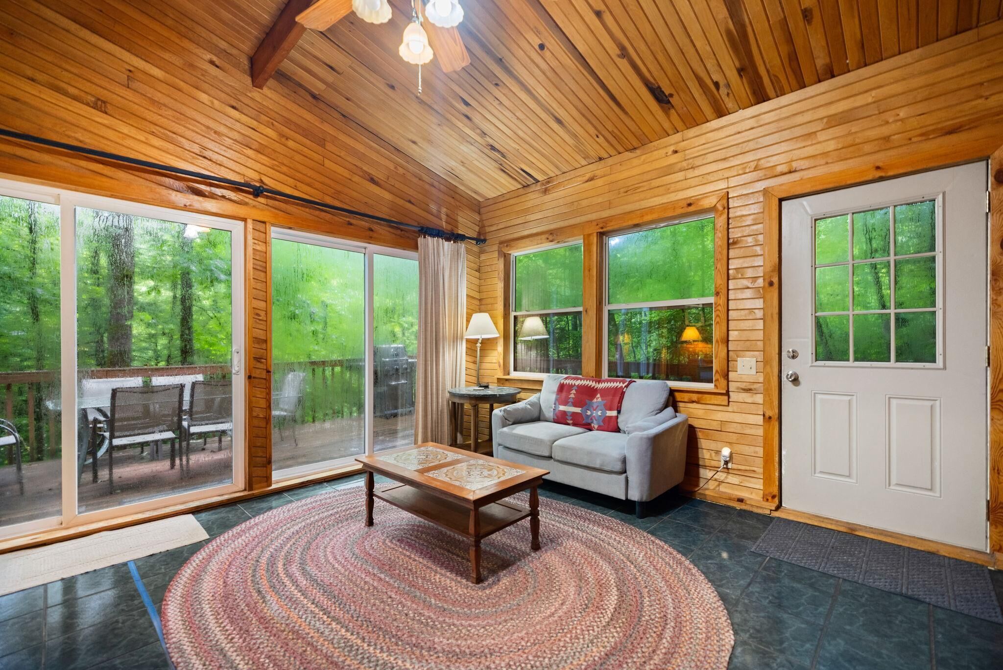 A cozy living room with a wooden ceiling, a sofa, a coffee table, and large windows offering a view of the outdoors.