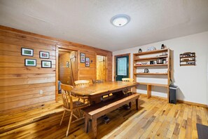 A wooden-paneled room with a dining table and chairs, a bench, and a shelf with kitchen items.