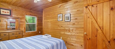 Une chambre à coucher aux murs lambrissés, avec un lit, un ventilateur de plafond, une fenêtre et deux tableaux encadrés accrochés au mur.