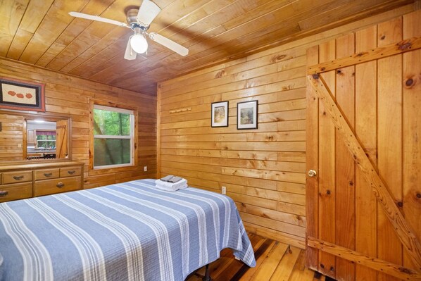 A wooden-paneled bedroom with a bed, a ceiling fan, a window, and two framed pictures on the wall.
