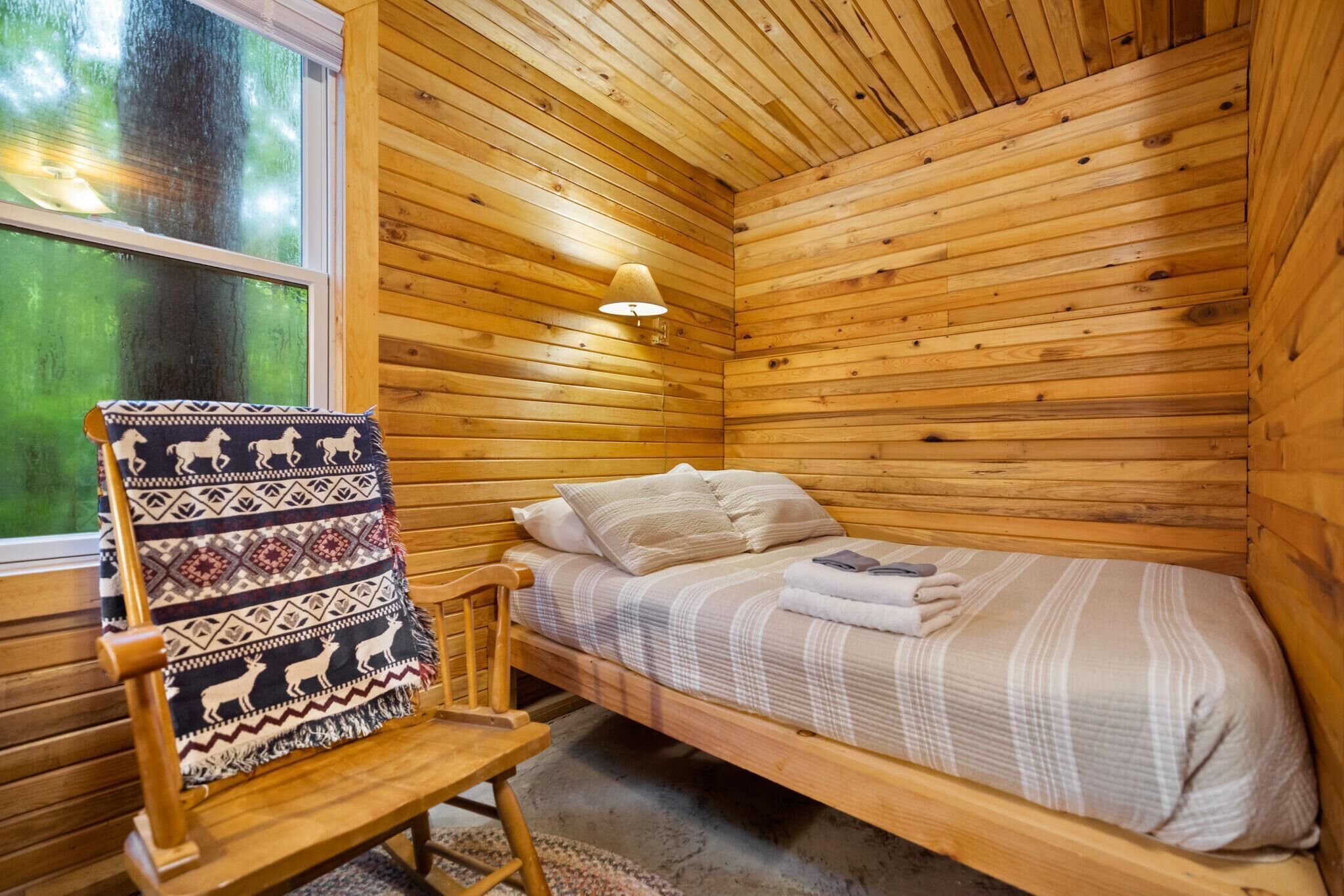 A wooden cabin room with a single bed, a chair with a patterned blanket, and a window with a view of greenery.