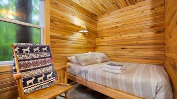 A wooden cabin room with a single bed, a chair with a patterned blanket, and a window with a view of greenery.