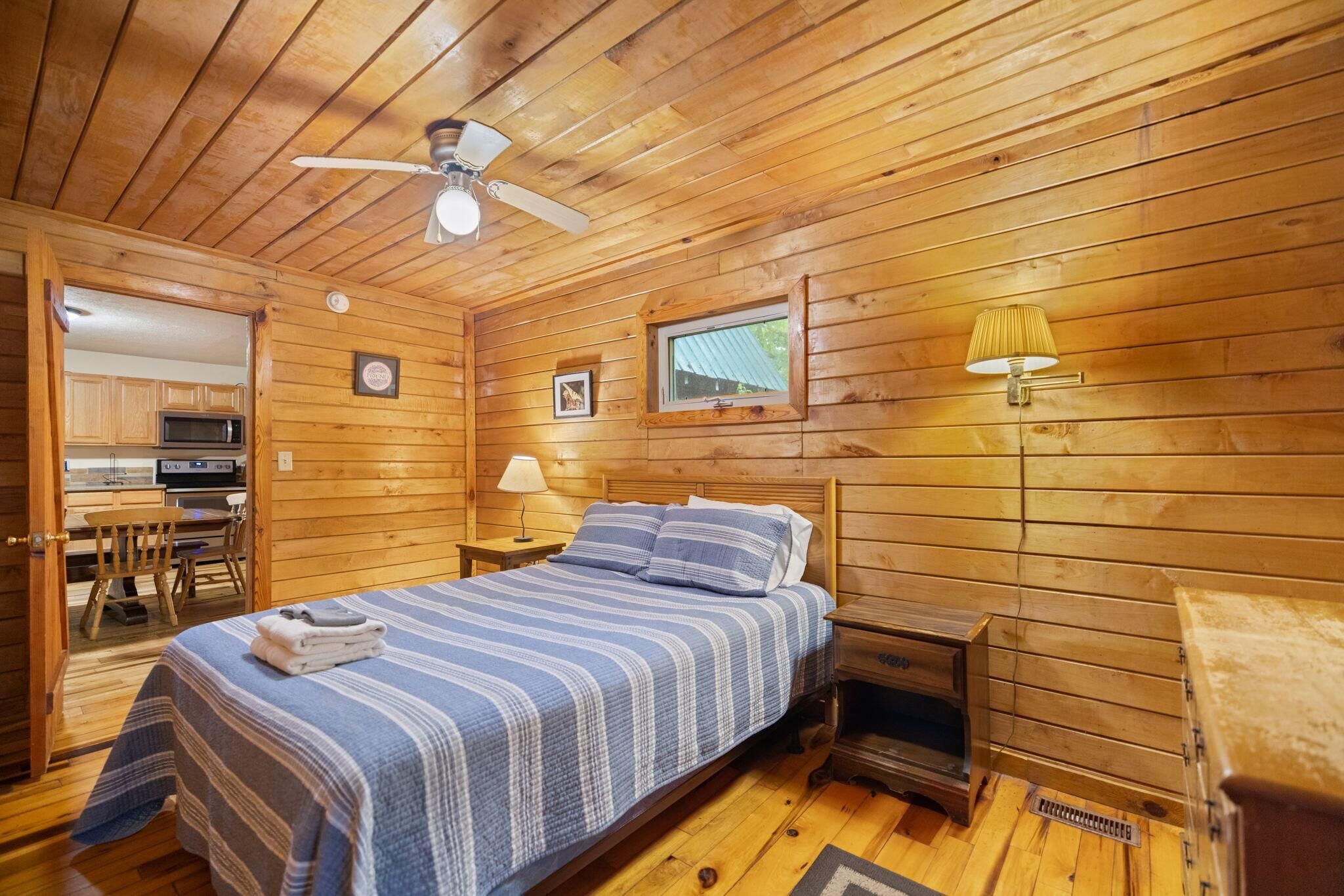 Un dormitorio tipo cabaña de madera con una cama, un ventilador de techo, una ventana y una pequeña cocina al fondo.