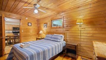 Ein Schlafzimmer einer Holzhütte mit einem Bett, einem Deckenventilator, einem Fenster und einer Küchenzeile im Hintergrund.