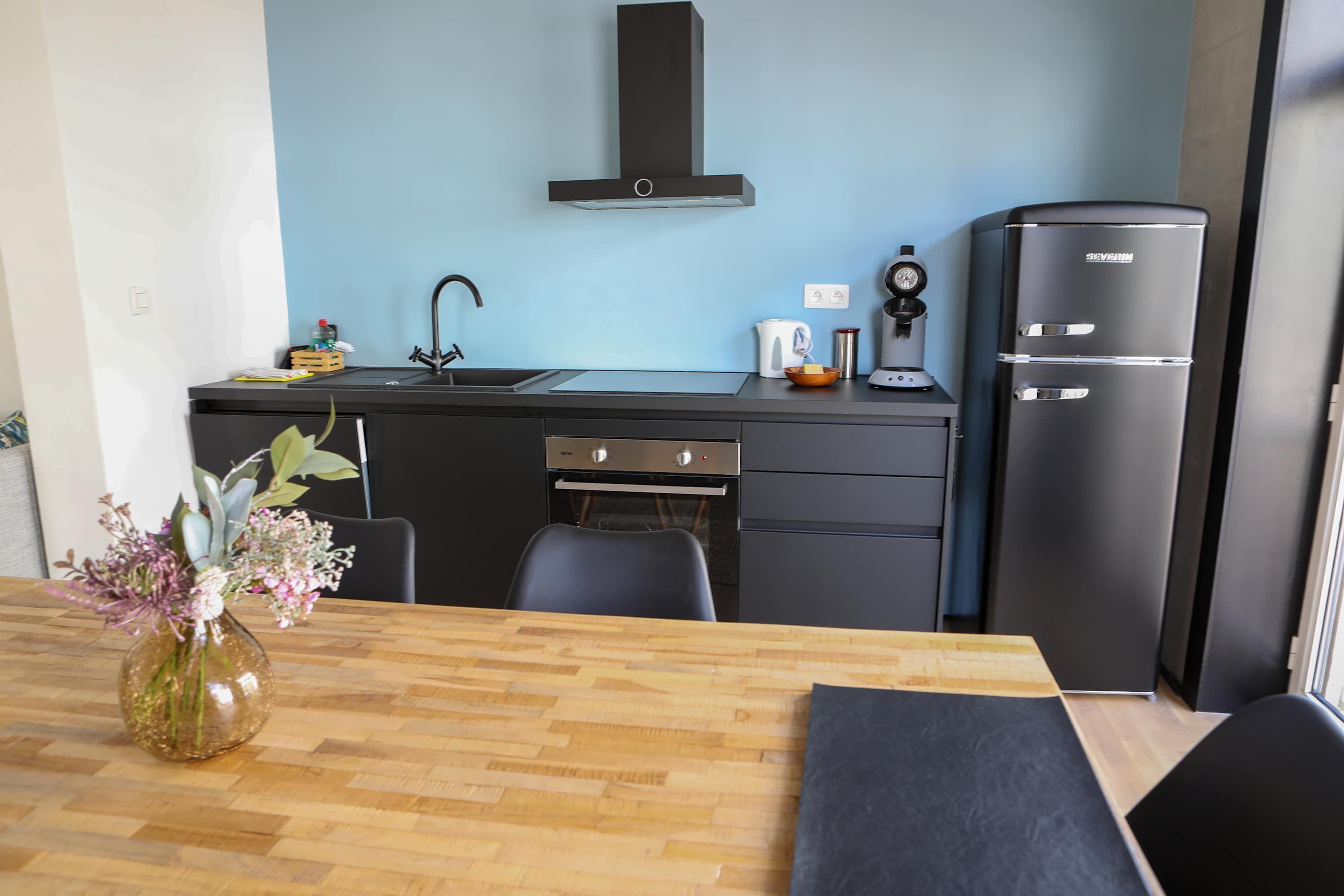 Een moderne keuken met een zwarte koelkast, een fornuis en een spoelbak. Er staat een houten tafel met een bloemenvaas.