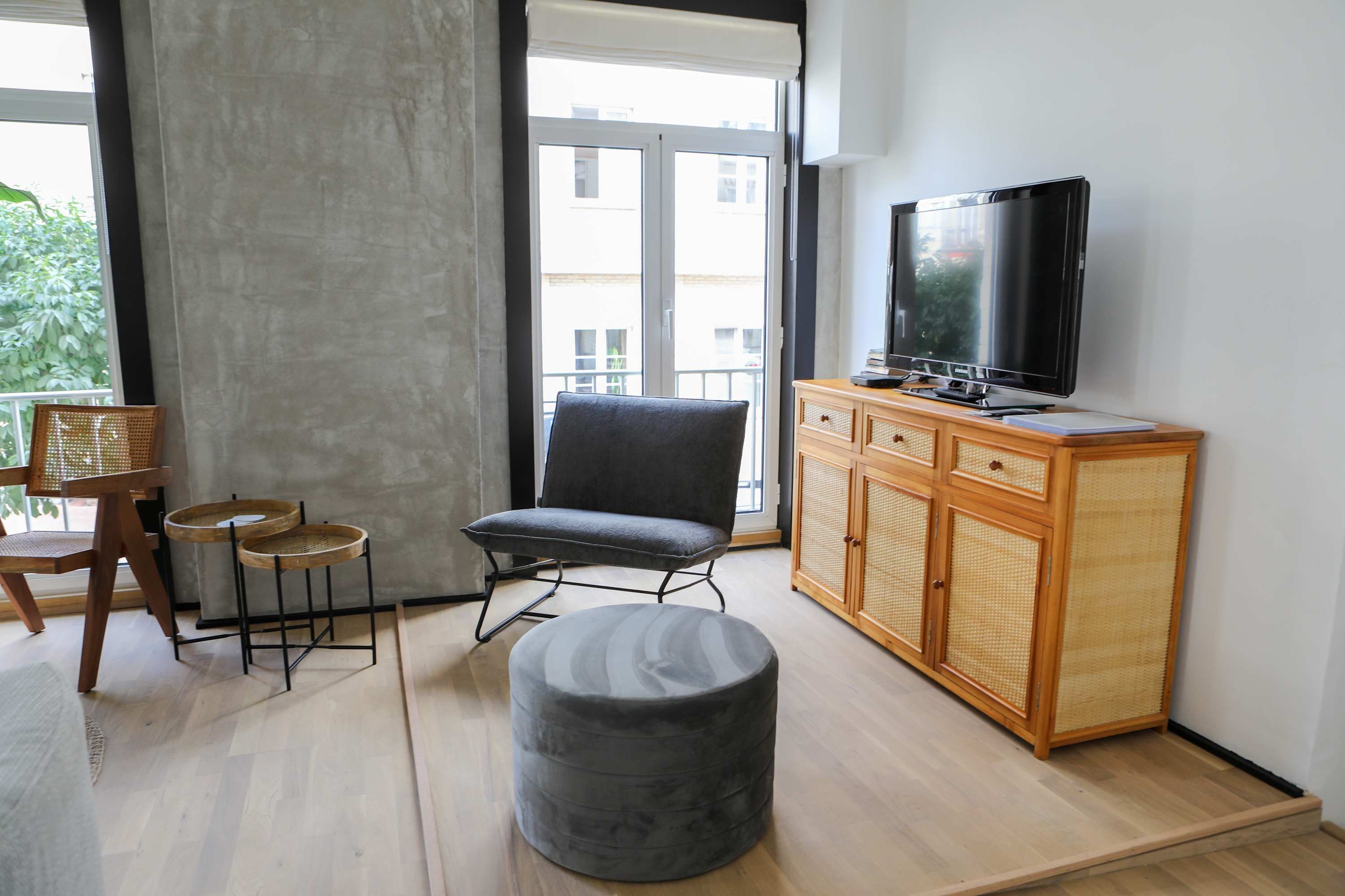 A modern living room with a grey armchair, a small round ottoman, a wooden cabinet with a television, and a wicker chair.