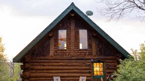 A log cabin with a green door, two windows, a deck with chairs, and a satellite dish. - Ely Log Cabin - Off Grid+Solar+WiFi - 40 acres to roam (Ely)