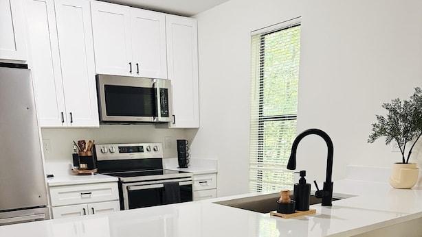 A modern kitchen with white cabinets, a large island with bar stools, a built-in microwave, and a window with blinds. - Charming Retreat in Raleigh, 2 bedrooms (Raleigh)