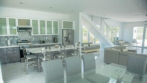 A modern kitchen with a central island, a dining area with a glass table, and a staircase leading to a balcony.