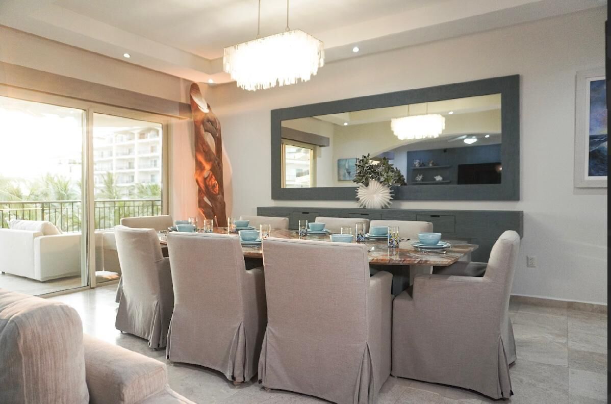A modern dining area with a large table set for six, surrounded by cushioned chairs. A large mirror and chandelier are mounted on the wall above the table.