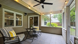 A covered patio with a ceiling fan, two chairs, a table, and large windows offering a view of the outdoors.