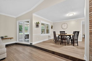 A dining area with a wooden table and chairs, a clock on the wall, and a sliding glass door leading to an outdoor space. - 3BR Retreat w/ Firepit + Big Garden | Washer/Dryer (Tallahassee)