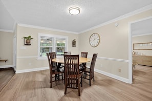 Un coin repas avec une table ronde et six chaises, une horloge au mur et une fenêtre avec des stores.