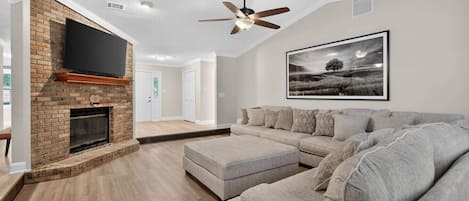 Un salon avec une cheminée, un ventilateur de plafond, un grand canapé et un tableau encadré au mur.