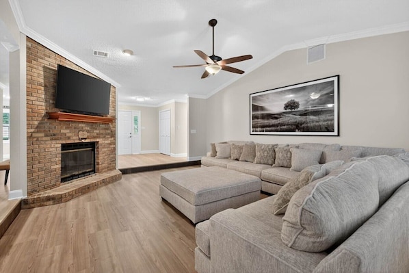 Un salon avec une cheminée, un ventilateur de plafond, un grand canapé et un tableau encadré au mur.
