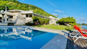 A poolside area with a clear view of the water, a wooden deck, and a building with a white exterior. - Le Ficus Grande Villa Bord de mer Avec Piscine (Paea)