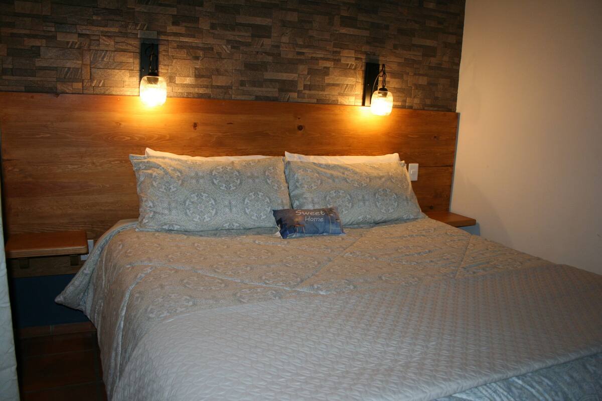 Una cama bien hecha con almohadas estampadas y un libro pequeño, un cabecero de madera y dos lámparas fijadas a la pared.