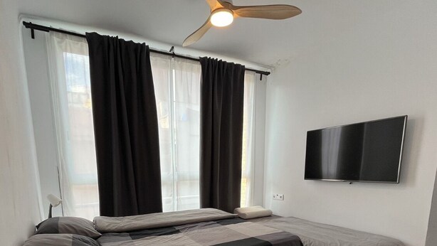 Een slaapkamer met een bed, een televisie, een plafondventilator en een raam met gordijnen.