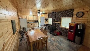 Interior de una cabaña de madera con una mesa de comedor, sillas, zona de cocina, pared de piedra y máquina de palomitas.
