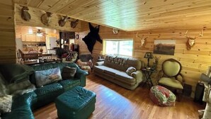 Interior rústico de una cabaña con paredes de madera, un sofá de cuero verde, un sofá beige y un comedor con armarios de madera.