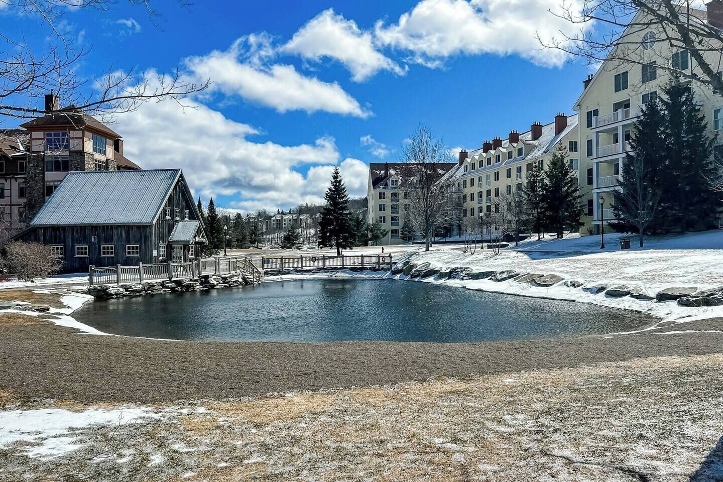 Blick auf einen See mit einer Holzsteganlage, einer Steinbrücke und Gebäuden im Hintergrund.