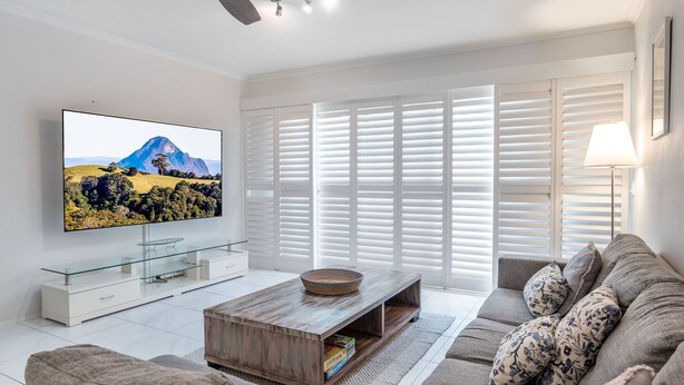 A modern living room with a flat-screen TV, a wooden coffee table, a sofa, and a large window with blinds.