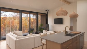 A modern kitchen with a large island, a dining area with a sofa, and a view of autumn trees. - The Ridge at Serà - Modern Retreat w/Sauna (Wardensville)