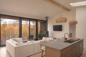 A modern kitchen with a large island, a dining area with a sofa, and a view of autumn trees.