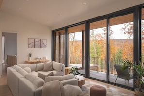 A modern living room with a sectional sofa, a coffee table, and a balcony with a view of autumn foliage.