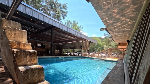 Une piscine avec des marches en pierre menant à l’eau, entourée d’une zone abritée dotée d’une terrasse en bois et offrant une vue sur les arbres et les collines en arrière-plan.