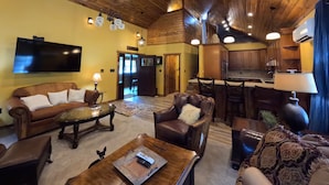 Un salon chaleureux avec un plafond en bois, un téléviseur à écran plat, des canapés en cuir et un coin repas donnant sur une cuisine en arrière-plan.