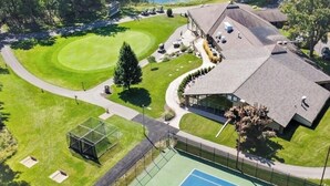 Vista aérea de un campo de golf con una cancha de tenis, un edificio de techo inclinado y un estanque.