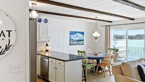 Una cocina moderna con armarios blancos, una isla central e iluminación colgante. Hay un comedor con mesa y sillas, y un sofá en primer plano.