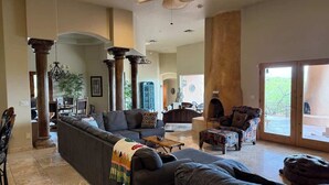Un salon spacieux avec un grand canapé d’angle, un fauteuil à motifs et une table basse. On y trouve également un ventilateur de plafond et une vue sur l’extérieur grâce à une porte-fenêtre coulissante.