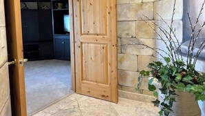 Un couloir aux murs de pierre, avec une porte en bois donnant sur une pièce. On peut voir une plante en pot sur la droite.