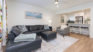 A modern living room with a sectional sofa, a grey armchair, and a small ottoman. There is a kitchen area with a refrigerator, a sink, and bar stools. A ceiling fan and recessed lighting are visible. - Newly Decorated BEAUTY in Downtown Ocean Springs with optional GOLF CART. (Ocean Springs)