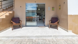 Un espacio al aire libre cubierto con dos sillas de madera, una entrada con puerta de vidrio, una escalera y piso de baldosas.