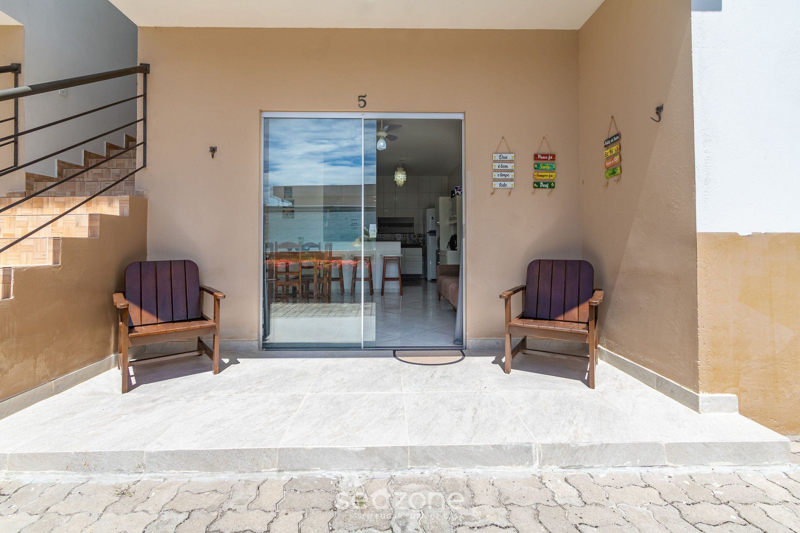 Un espacio al aire libre cubierto con dos sillas de madera, una entrada con puerta de vidrio, una escalera y piso de baldosas.