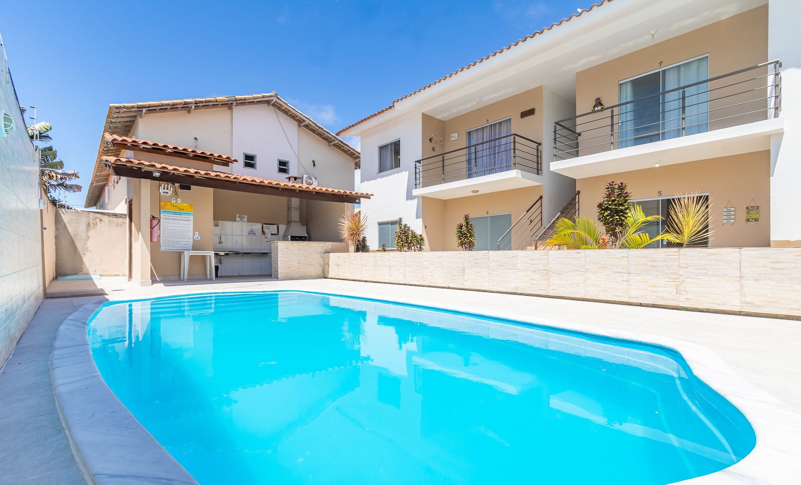 Una piscina con una zona de cocina al aire libre techada, un edificio de dos pisos con balcones, y un cielo azul despejado.