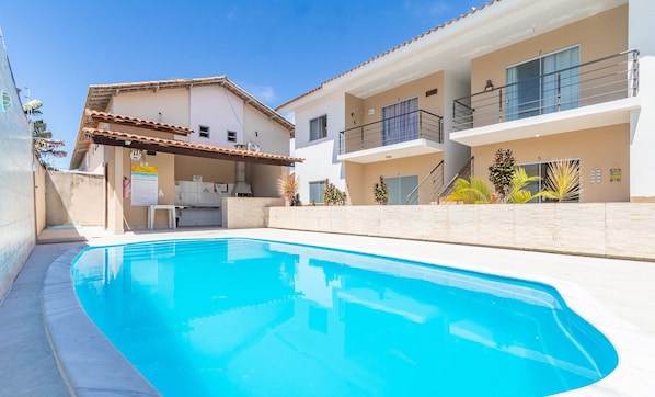 Una piscina con una zona de cocina al aire libre techada, un edificio de dos pisos con balcones, y un cielo azul despejado.