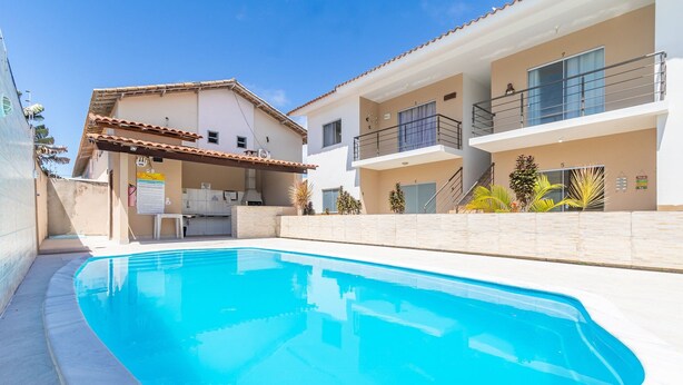 Una piscina con una zona de cocina al aire libre techada, un edificio de dos pisos con balcones, y un cielo azul despejado.