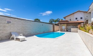 A swimming pool with a tiled deck, a white lounge chair, and a small covered area with a sink.