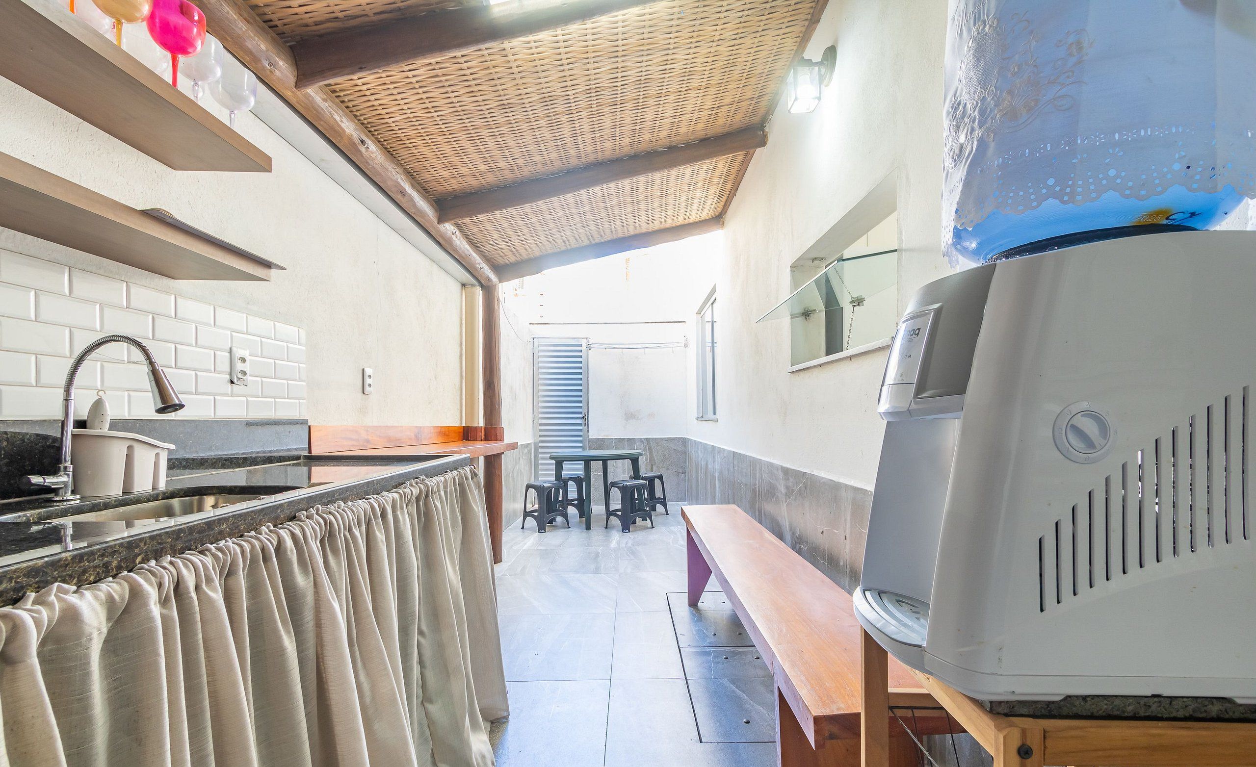 Une cuisine avec un mur recouvert de carreaux, un banc en bois, un barbecue et un coin repas avec des tabourets.