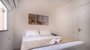 A neatly made bed with a beige blanket and rolled towels, a bedside table with a lamp, and abstract wall art above the bed. - PDC - Apartments in a Condominium With a Pool in Ilheus (Ilhéus)