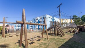 A playground with swings and climbing structures, surrounded by a fence, with residential buildings in the background. - PDC - Apartments in a Condominium With a Pool in Ilheus (Ilhéus)
