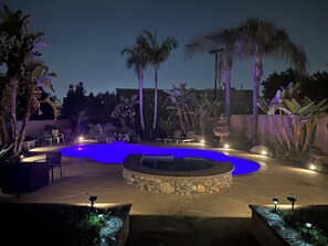 A circular pool with a stone edge, surrounded by palm trees and chairs, with a stone wall in the background.