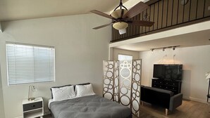 Ein Schlafzimmer mit Bett, Fernseher, Deckenventilator, Nachttisch und Fenster mit Jalousien.