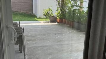 On aperçoit, à travers une porte vitrée, un espace extérieur aménagé avec un paillasson, un banc et des plantes en pot.